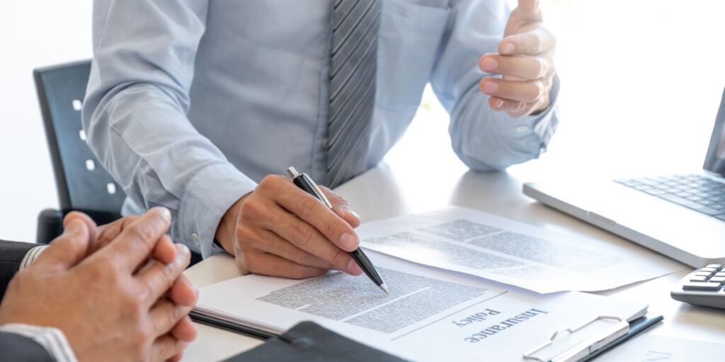 Two professionals in a business setting discussing an insurance policy document. One person is holding a pen, gesturing toward the paperwork on the table, while a laptop and calculator are nearby.